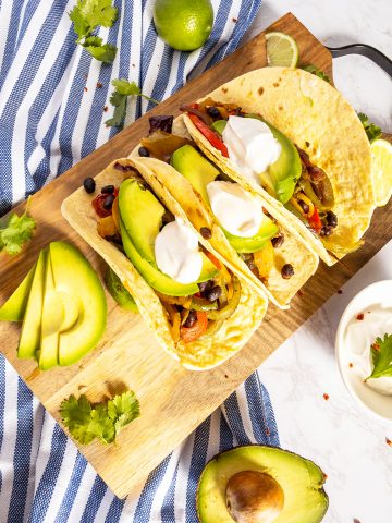 three vegetarian fajitas with sour ceeam and avocado slices on a wooden chopping board