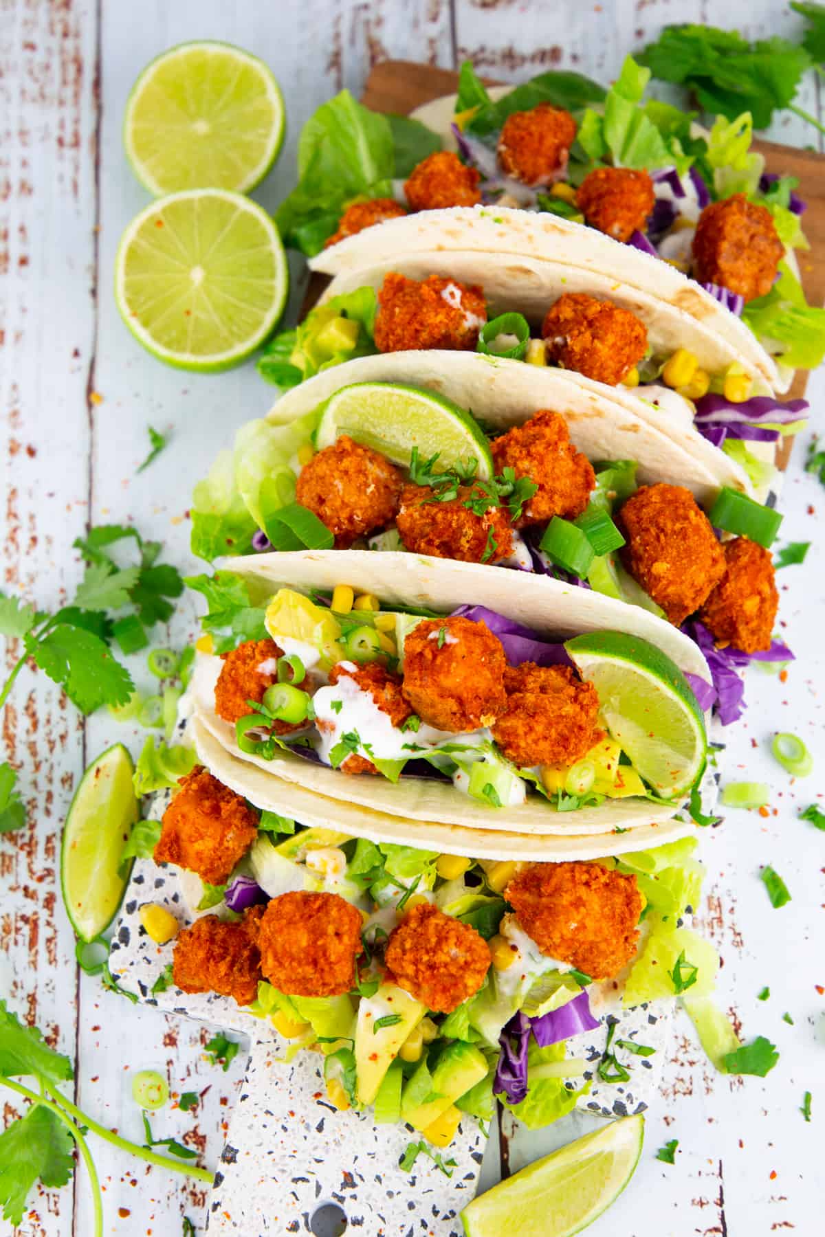 tacos filled with baked tofu cubes, lettuce, and avocado on a wooden board with limes on the side