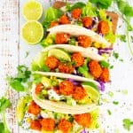 tacos filled with baked tofu cubes, lettuce, and avocado on a wooden board with limes on the side
