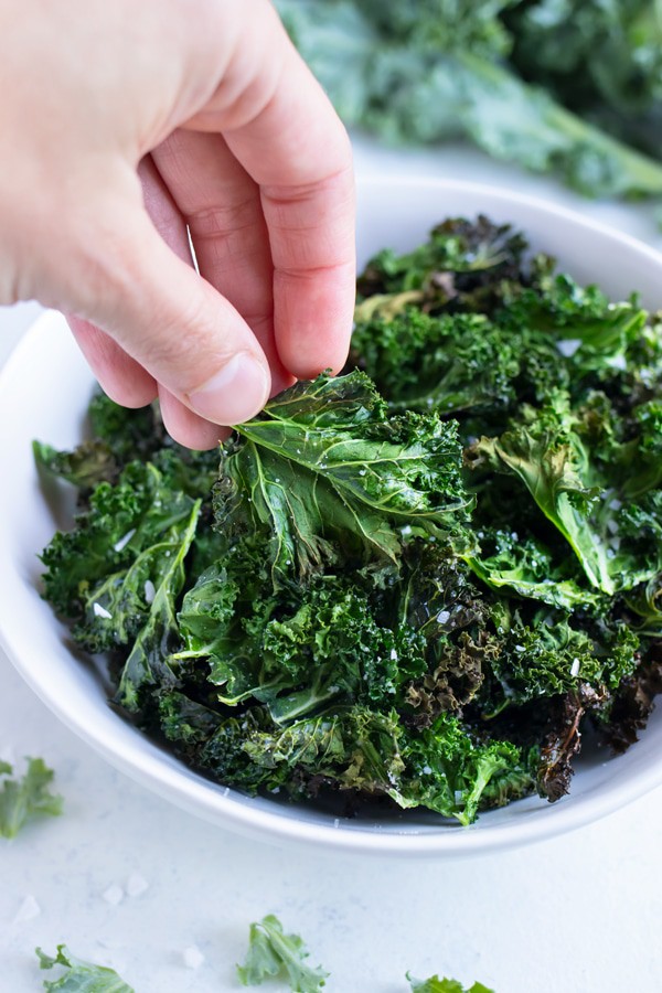 a hand grabbing kale chips out of a white bowl 