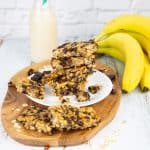 a stack of vegan granola bars on a white plate with bananas and a bottle of milk in the background