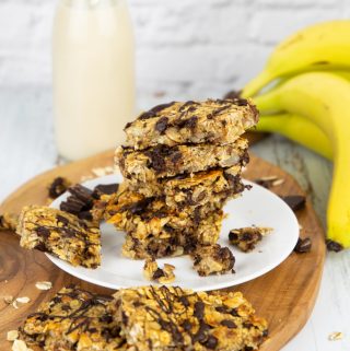 a stack of vegan granola bars on a white plate with bananas and a bottle of milk in the background
