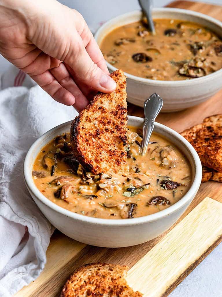 a hand dipping a slice of bread in a bowl of vegan mushroom soup 