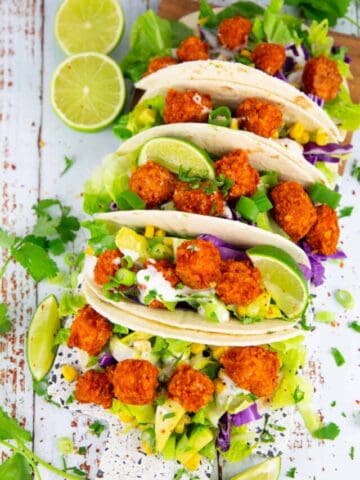 tacos filled with baked tofu cubes, lettuce, and avocado on a wooden board with limes on the side