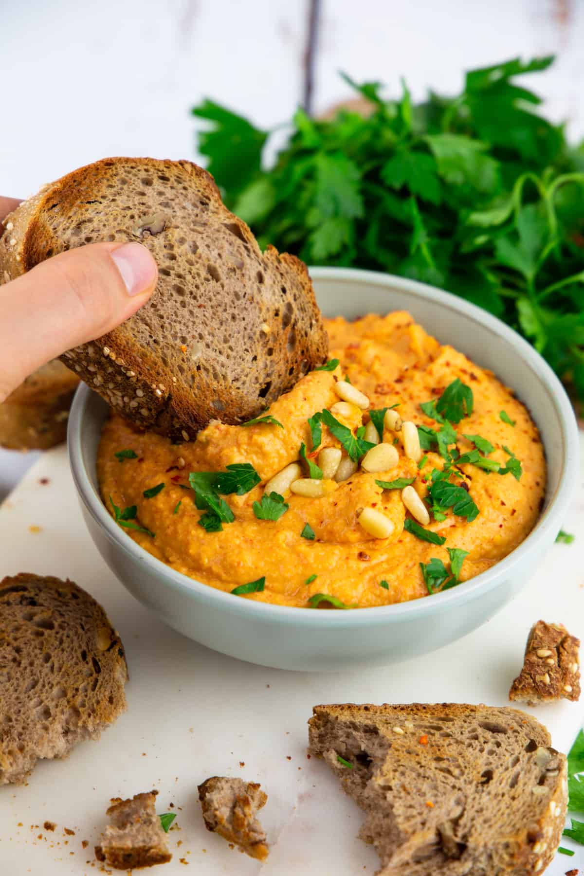a hand dipping a piece of bread into a bowl of roasted red pepper hummus