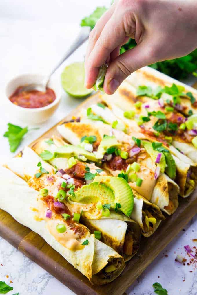 taquitos on a wooden board covered with chipotle mayo, avocado, and green onions