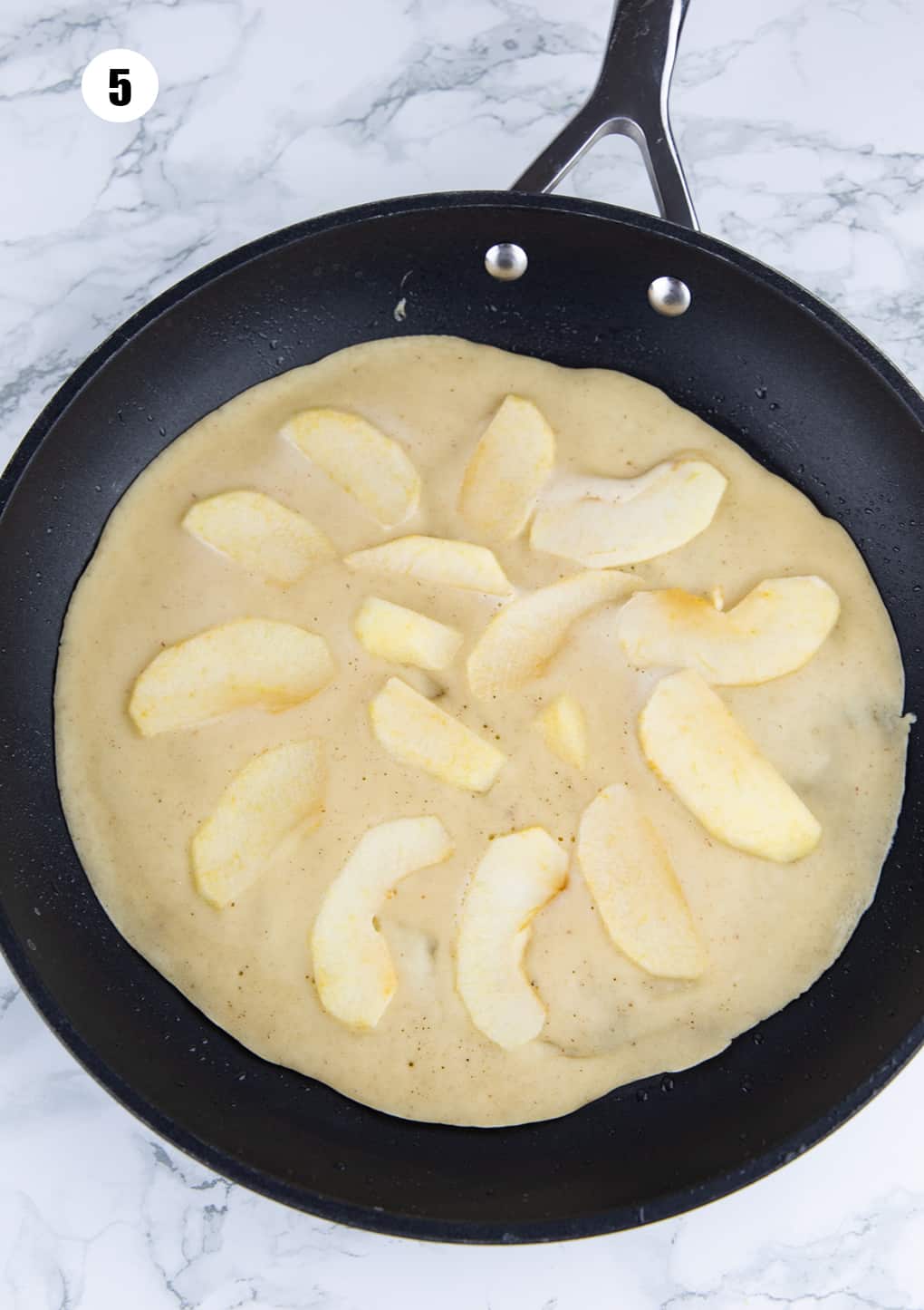 a pancake topped with apple slices in a large pan on a marble countertop 
