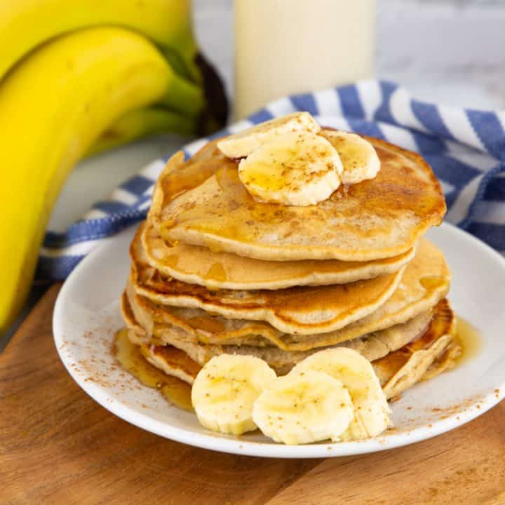 a stack of vegan banana pancakes on a white plate with bananas in the background