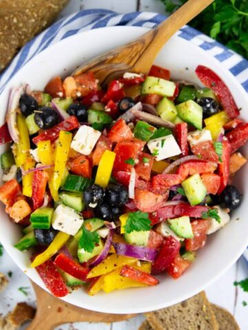 a salad with tomatoes, cucumber, and bell pepper in a white bowl with a wooden spoon