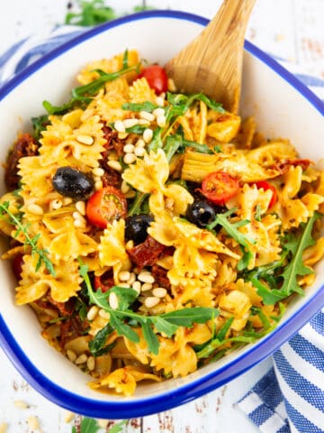 Pasta salad in a white bowl with arugula and pine nuts on the side