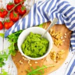 arugula pesto in a small white bowl on a wooden chopping board with tomatoes in the background