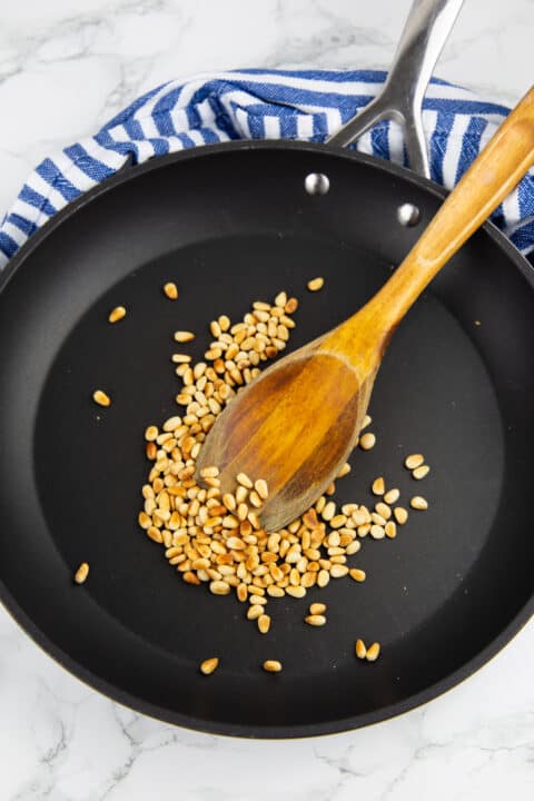 roasted pine nuts in a black pan with a wooden spoon on a marble countertop