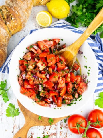 a white bowl with tomato salad with a wooden spoon with a loaf of bread in the background