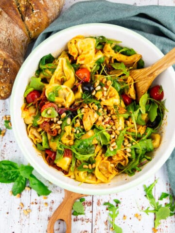 a white bowl with tortellini salad with a wooden spoon with bread in the background