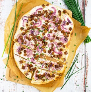 a Flammkuchen on a wooden chopping board lined with parchment paper with chives in the background
