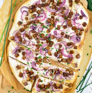 a Flammkuchen on a wooden chopping board lined with parchment paper with chives in the background