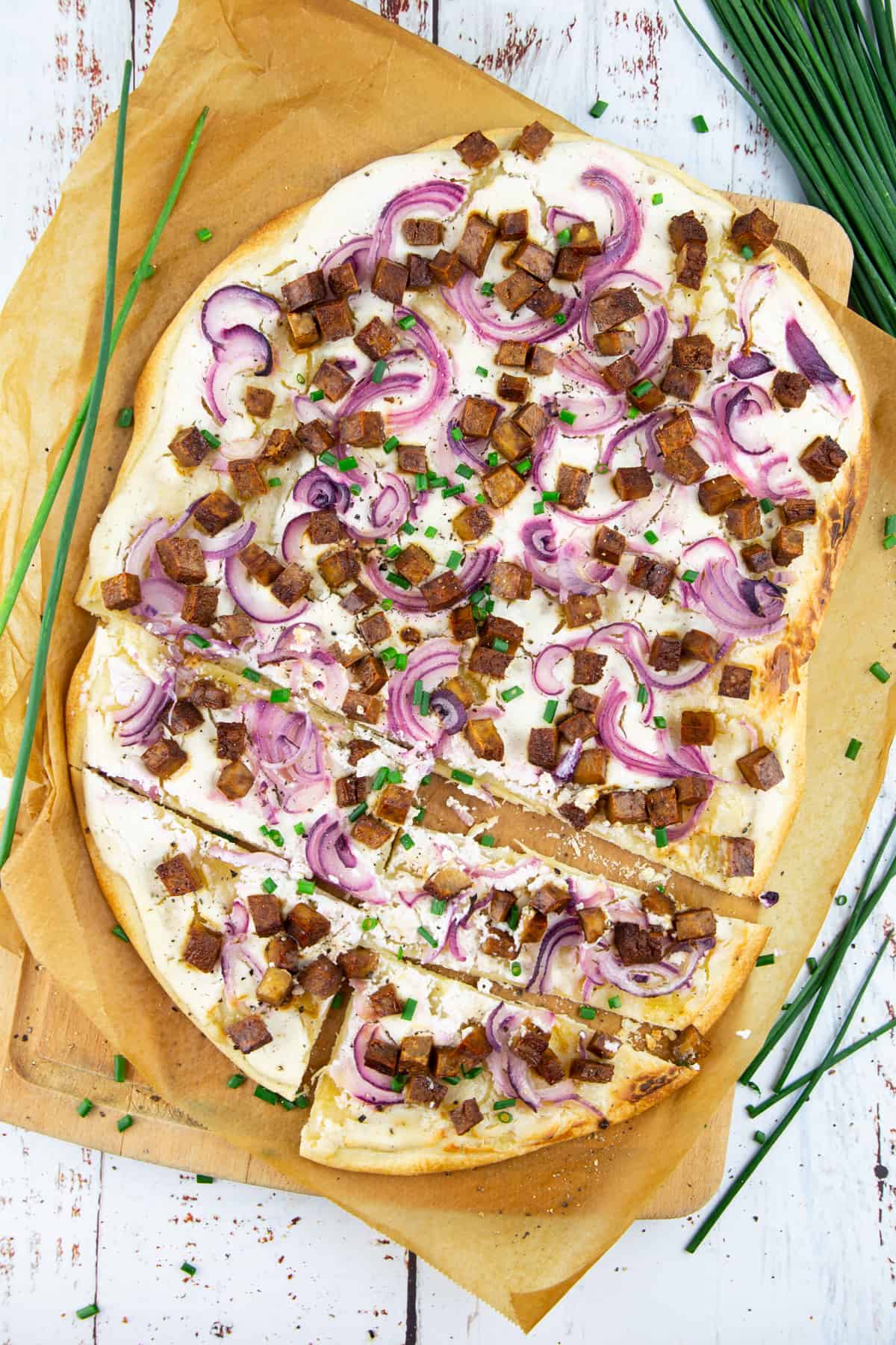 a Flammkuchen on a wooden chopping board lined with parchment paper with chives in the background 