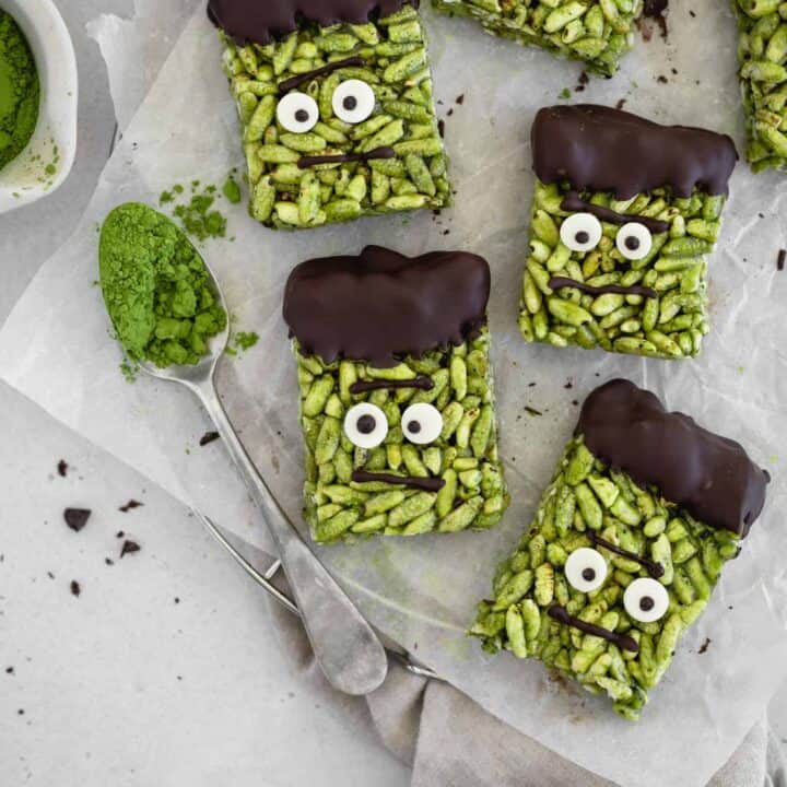 five Frankenstein rice crispy treats on a sheet of baking paper with a spoon with matcha powder on the side