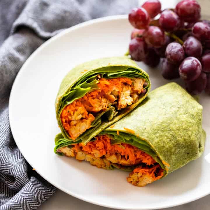 two buffalo tempeh wraps on a white plate with red grapes on the side
