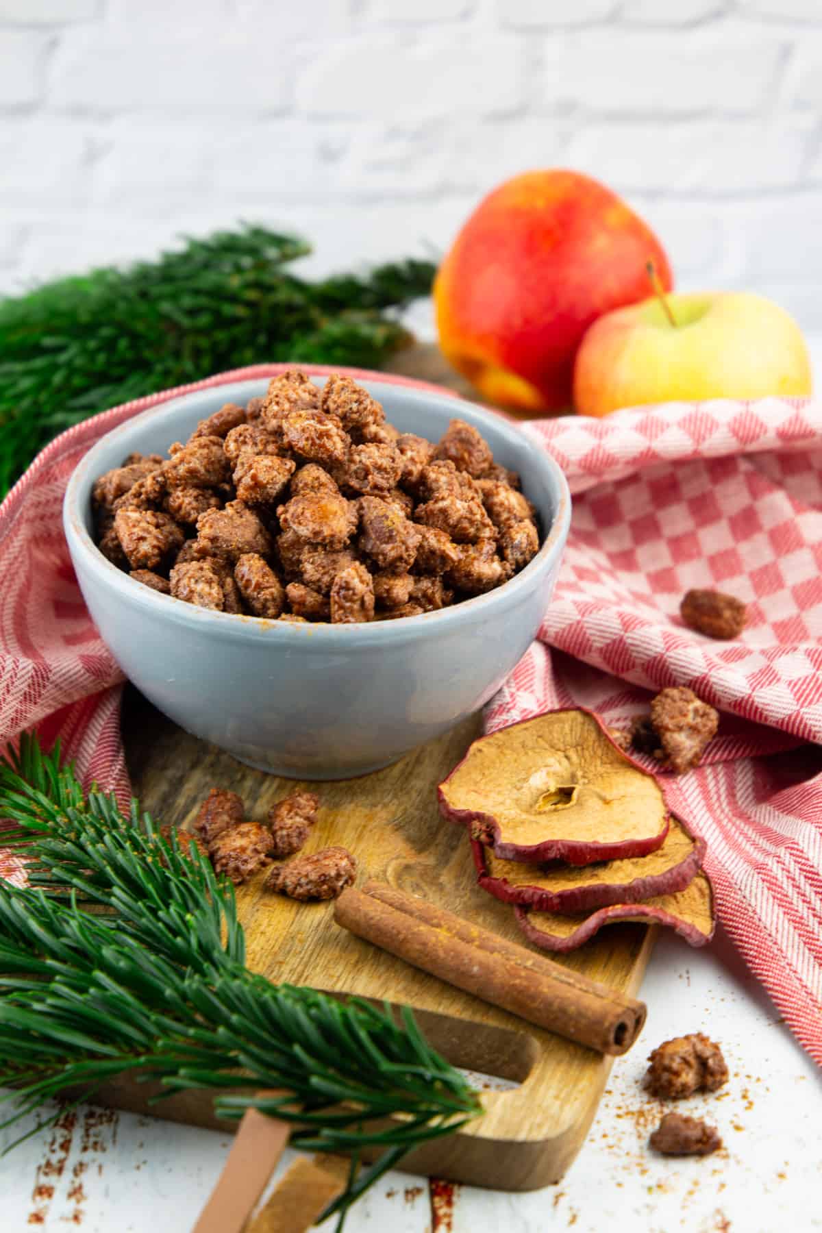 a small grey bowl of candied almonds on a wooden board with two apples and evergreens in the background 