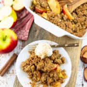 vegan apple crumble in a white casserole dish on a wooden board with a plate on the side