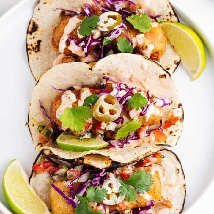 three vegan fish tacos on a white plate with lime wedges on the side