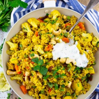 vegetable biryani in a white pan on a wooden board with parsley and mint in the background