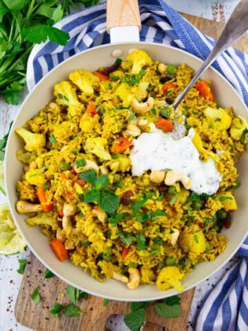 vegetable biryani in a white pan on a wooden board with parsley and mint in the background