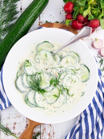 a white bowl with dill cucumber salad with radishes and a cucumber in the background