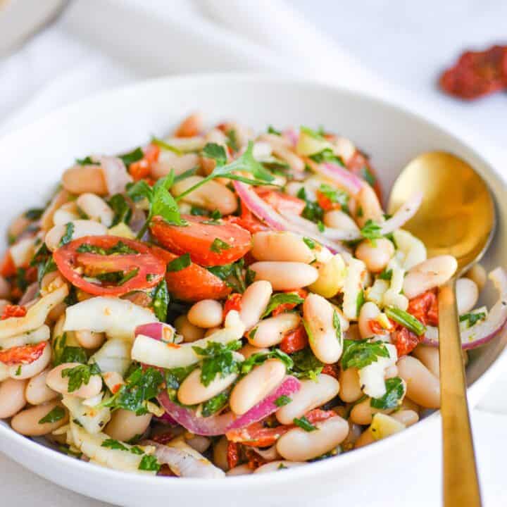 a white bowl with Tuscan bean salad with a golden spoon on a white countertop