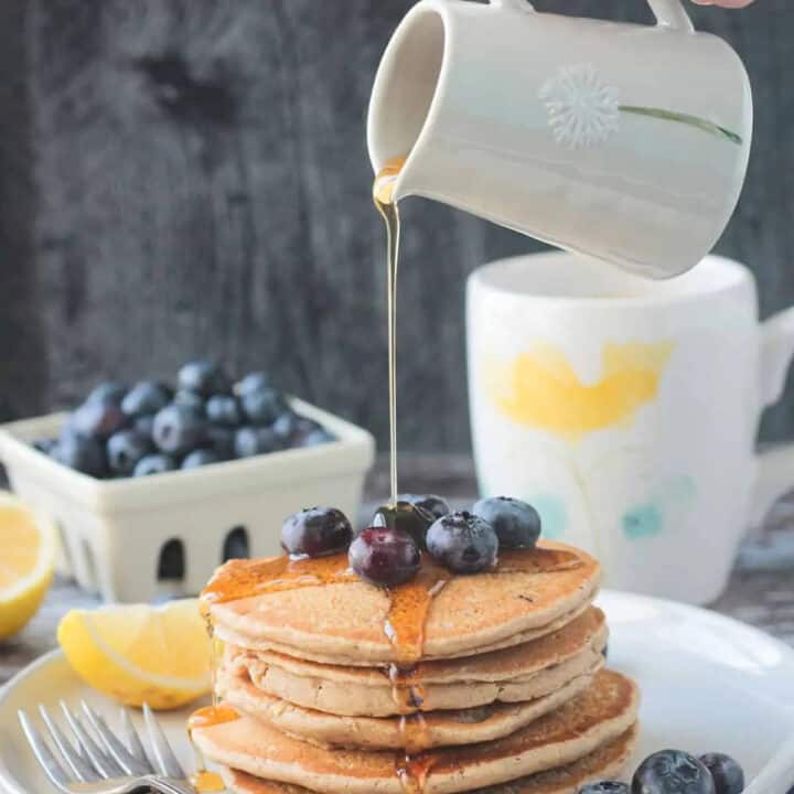 a hand pouring maple syrup over a stack of vegan oat pancakes with a little white jug