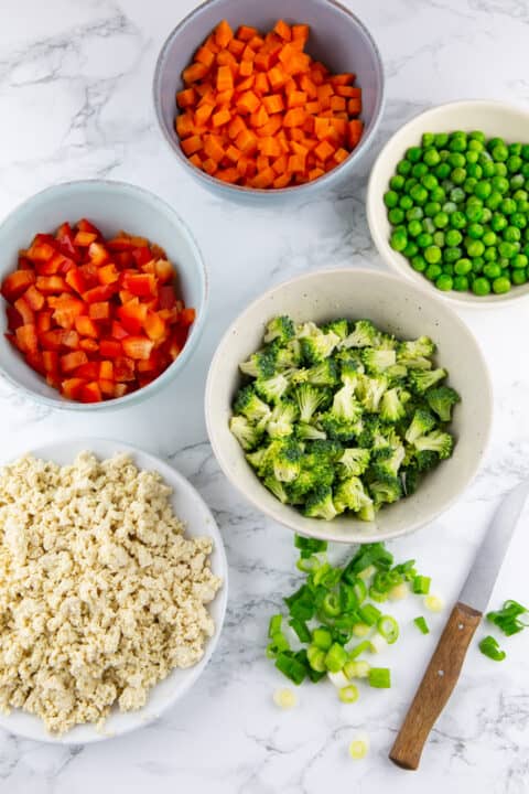 chopped vegetables, peas, and crumbled tofu in small bowls on a wooden countertop