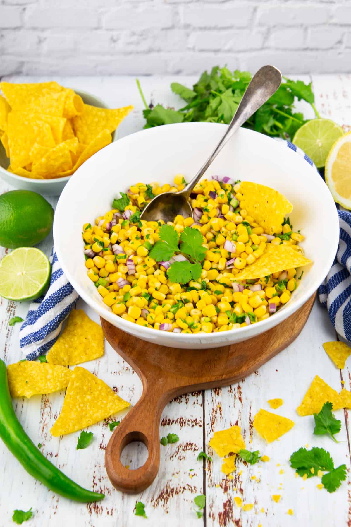 a white bowl with corn salsa with a spoon on a white wooden board with nachos and cilantro in the background