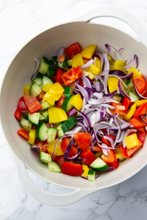 cucumber, bell pepper, tomatoes, and red onion in a grey bowl on a marble countertop