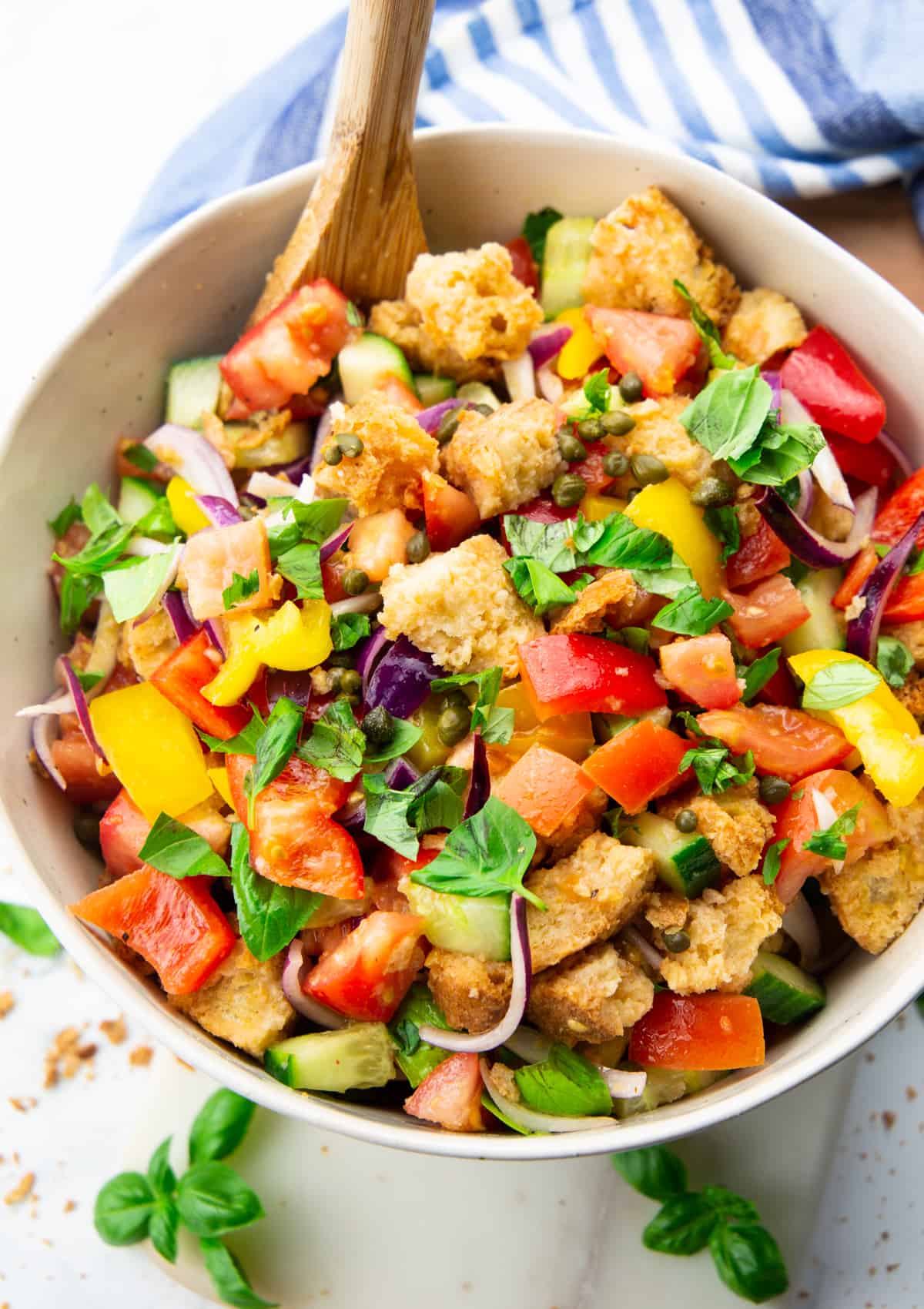 Panzanella in a beige bowl with a wooden spoon on a marble countertop with basil on the side