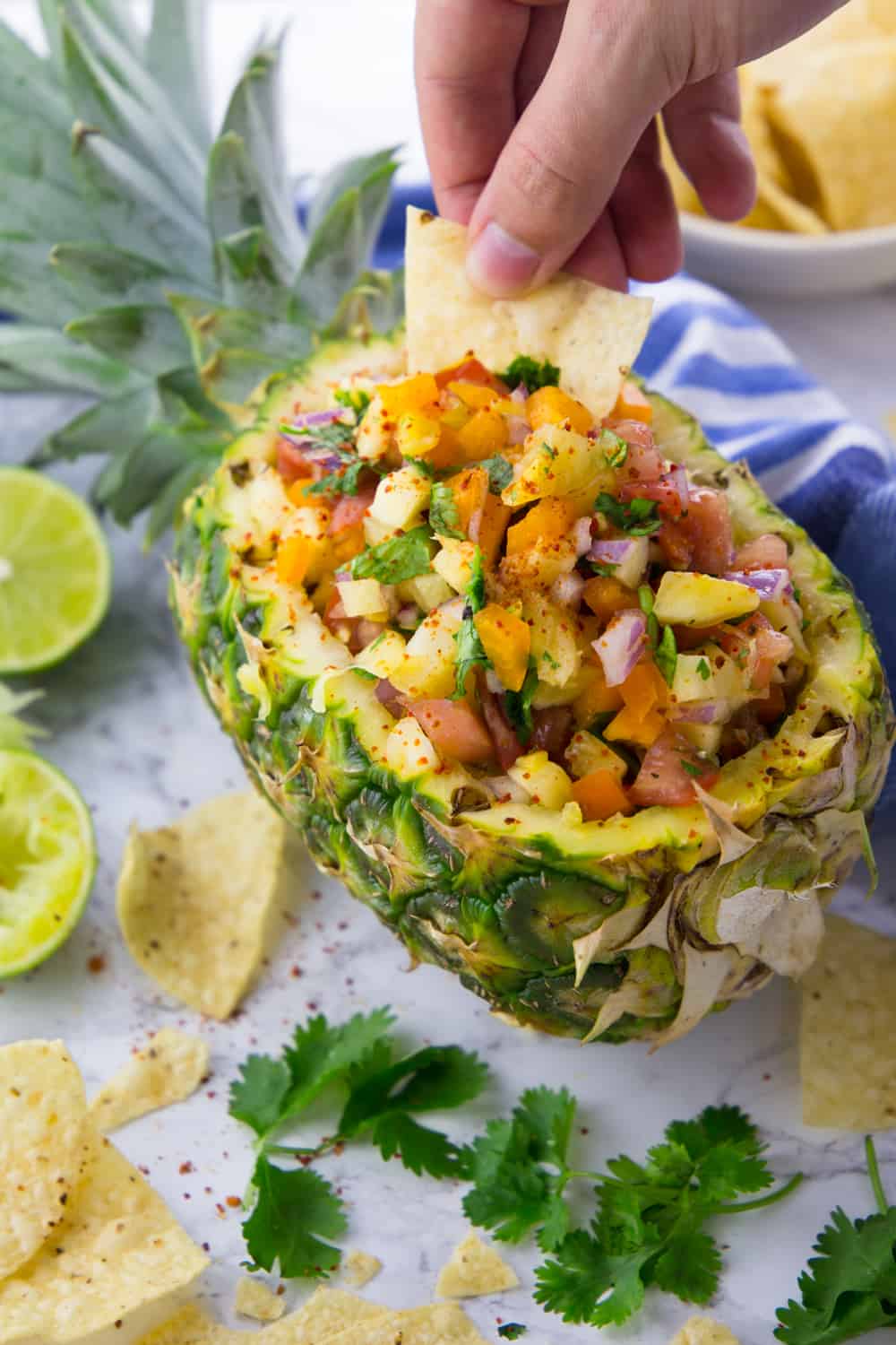 a hand dipping a tortilla chip into a halved pineapple filled with pineapple salsa