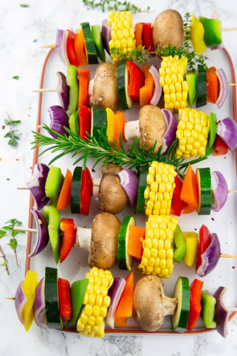 uncooked vegetable skewers with fresh herbs on a white plate