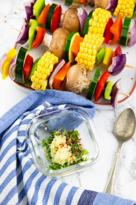 vegetable kabobs on a white plate with a small bowl with chopped herbs and garlic on the side