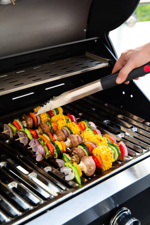 six vegetable kabobs on a grill with a hand holding barbecue tongs