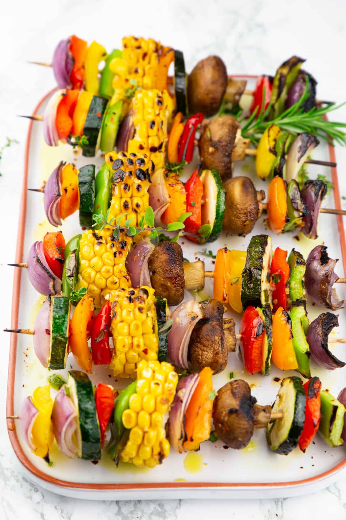 six vegetable kabobs with fresh rosemary on a white plate on a marble countertop 