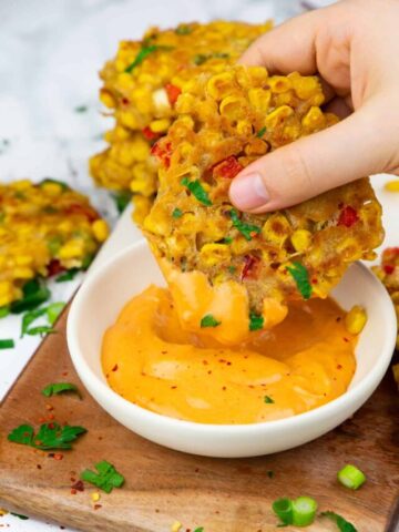 a hand dipping a corn fritter into a small bowl with spicy mayonnaise on a wooden board
