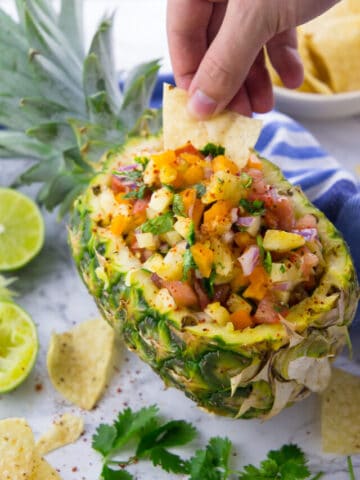 a hand dipping a tortilla chip into a halved pineapple filled with pineapple salsa