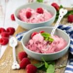 two bowls with raspberry ice cream on a wooden board with fresh raspberries on the side