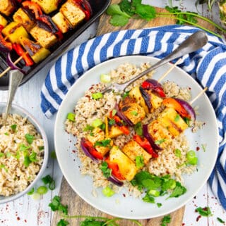 two tofu vegetable skewers on a grey plate with brown rice with a pan with more skewers in the background