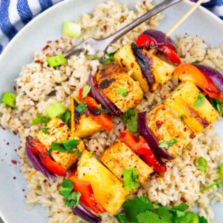 two tofu vegetables skewers with pineapple and red bell pepper on a plate with brown rice