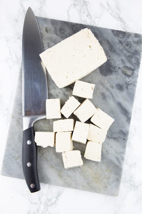 tofu cubes on a chopping board on a marble countertop with a knife on the side