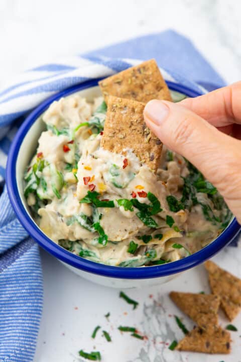 a hand dipping a cracker in a bowl with spinach artichoke dip
