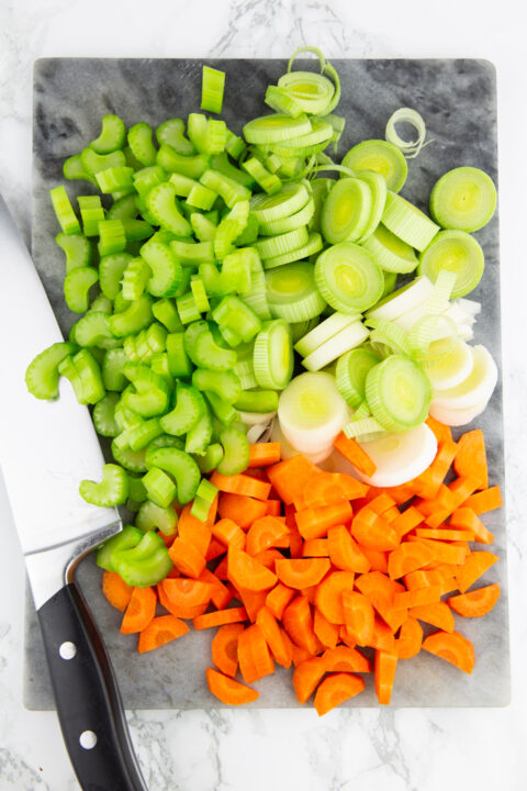 chopped celery, leek, and carrots on a marble countertop with a large knife