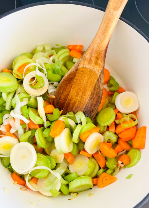 leek, carrots, and celery in a white pot with a wooden spoon