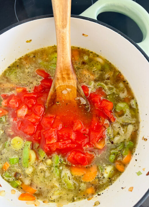 a white pot with vegetables, vegetable broth, and diced tomatoes with a wooden spoon